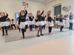 Dziewczynki ubrane na czarno w białych spódniczkach, stoją na jednej nodze, na szarym dywanie i ruszają rękami. Pokazują taniec.