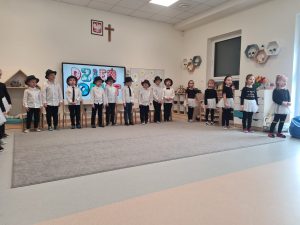 Chłopcy i dziewczynki ubrani na biało czarno stoją na około szarego dywanu i śpiewają piosenkę.