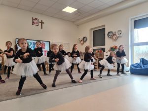 Dziewczynki ubrane na czarno w białych spódniczkach, stoją w rozkroku na szarym dywanie i ruszają rękami. Pokazują taniec.
