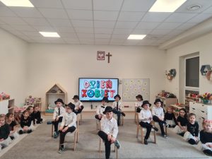 Chłopcy ubrani w białe koszule, siedzą na krzesłach i mają opartą nogę na nodze. Na głowach mają czarne kapelusze. Chłopcy pokazują taniec.