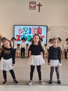 Trzy dziewczynki ubrane na czarno w białych spódniczkach stoją na szarym dywanie i śpiewają piosenkę.