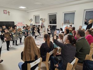 Rodzice siedzą na krzesłach oglądają występujące dzieci przebrane za koty.
