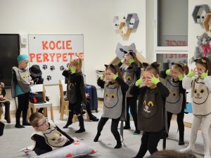 Dziewczynki ubrane na czarno w kamizelkach z wizerunkiem kota, stoją na szarym dywanie i tańczą do muzyki. Na poduszce siedzi jedno dziecko ubrane w strój kotka.