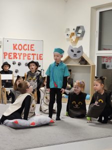 Chłopiec przebrany za doktora stoi przed poduszką na której siedzi chłopiec ubrany w strój kota. Z boku szarego dywanu siedzą dziewczynki w strojach kotów.