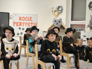 Chłopcy ubrane w strój kota, siedzą na krzesłach i mają założoną nogę na nogę. Na głowach mają kapelusze.