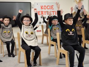 Chłopcy ubrane w strój kota, siedzą na krzesłach i podnoszą ręce do góry nad głowę.