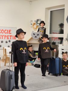 Dwoje chłopców stoi na dywanie obok walizki, ubrani są w strój kota. Jeden chłopiec w cylindrze stoi wysoko za domkiem.