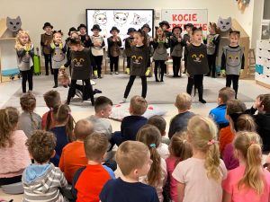 Dzieci siedzą na szarej podłodze i patrzą na dzieci występujące w strojach kotów.