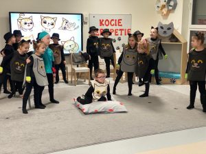 Dzieci poubierane w stroje kotów stoją w parach i trzymają się za ramiona. Pośrodku na poduszce siedzi dziecko w stroju kota.