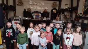 Na szarej ścianie widnieje napis Muzeum Piekarnictwa. Pod napisem na kredensie stoją stare eksponaty. zdjęcia rodziny Vogt. Przed tym kredensem stoi grupa dzieci, która pozuje do zdjęcia.