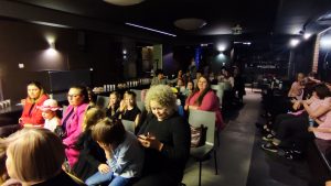 Dzieci i osoby dorosłe siedzą na krzesłach i patrzą na scenę czarną.