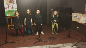 Cztery dziewczynki w czarnych strojach ze skrzydłami w kolorach czerwonym, niebieskim i żółtym. Stoją na scenie i śpiewają piosenkę do mikrofonów.
