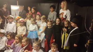 Bardzo duża grupa dzieci stoi obok siebie, ubrani kolorowo. Za nimi stoją osoby dorosłe - jury konkursu wokalnego.