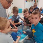 Chłopcy siedząc z panią, przy pojemniku wypełnionym niebieską masą sensoryczną szukają elementów, aby zrobić z nich łancuch.