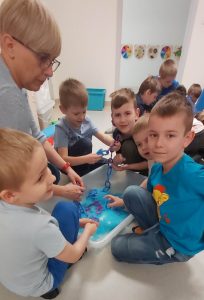 Chłopcy siedząc z panią, przy pojemniku wypełnionym niebieską masą sensoryczną szukają elementów, aby zrobić z nich łancuch.