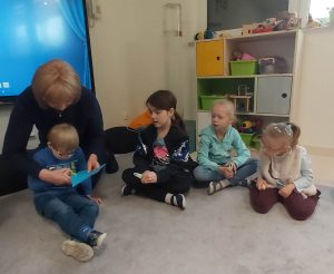 Dzieci siedzą w kole. Podają sobie niebieskie papierowe serce, za pomocą klamerek trzymanych w rączkach.