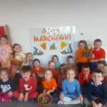 ZDJĘCIE GRUPOWE NA TLE TABLICY Z NAPISEM DZIEŃ MARCHEWKI