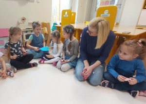 Dzieci siedzą w kole. Podają sobie niebieskie papierowe serce, za pomocą klamerek trzymanych w rączkach.