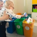 Dziewczynka z pomocą nauczyciela umieszcza śmieci w odpowiednim koszu.