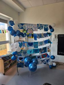 Tablica, na niej wiszą prace plastyczne, na których są niebieskie serduszka. Obok niebiesko-białe balony.