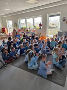 Dzieci ubrane na niebiesko siedzą na dywanie i patrzą w stronę dużego ekranu.