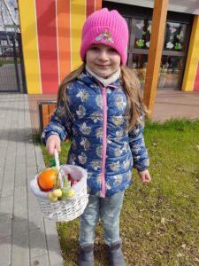 Dziewczynka w kolorowej kurtce i różowej czapce trzyma w ręce koszyczek wielkanocny. Stoi na placu zabaw.