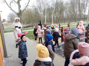 Duża grupa dzieci ubranych w kurtki i czapki są na placu zabaw i szukają koszyczków wielkanocnych. Obok nich stoi biały zajączek.