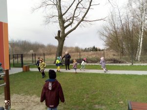 Grupka dzieci ubranych w kurtki i czapki biega po placu zabaw i szuka wielkanocnych koszyczków.