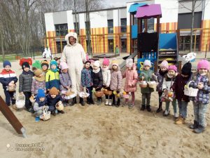 Duża grupa dzieci z koszyczkami wielkanocnymi stoją obok białego zajączka na placu zabaw.