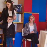 Chłopiec w okularach w białej koszuli i granatowym swetrze stoi na tle regałów z koszykami. W ręce trzyma palmę wielkanocną, którą wręcza pani.