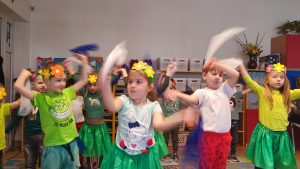 Dzieci ubrane na kolorowo, na głowach wianki w kwiaty, machają białymi chustkami do muzyki.