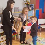 Chłopiec w okularach w białej koszuli i granatowym swetrze, odbiera nagrodę za udział w konkursie z rąk pani w czarnej marynarce i pani w szarej marynarce.