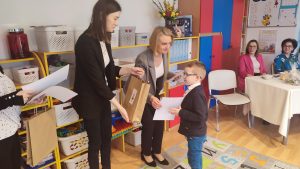 Chłopiec w okularach w białej koszuli i granatowym swetrze, odbiera nagrodę za udział w konkursie z rąk pani w czarnej marynarce i pani w szarej marynarce.