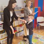 Chłopiec w okularach w białej koszuli i granatowym swetrze, odbiera nagrodę za udział w konkursie z rąk pani w czarnej marynarce.
