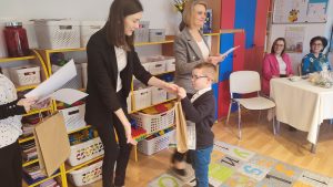 Chłopiec w okularach w białej koszuli i granatowym swetrze, odbiera nagrodę za udział w konkursie z rąk pani w czarnej marynarce.