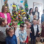 Dzieci i osoby dorosłe stoją na tle palm wielkanocnych.