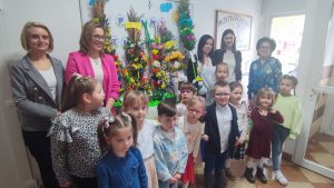 Dzieci i osoby dorosłe stoją na tle palm wielkanocnych.