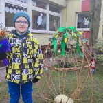 Chłopiec ubrany w kurtkę zielono- żółtą, w niebieskiej czapce w okularach, w ręce trzyma palmę wielkanocną. Stoi obok wiklinowego jajka.