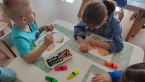 Dzieci siedzą przy stole białym i malują pisakami i kredkami papierową pisankę.