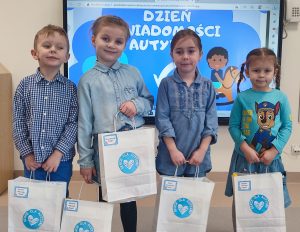 Trzy dziewczynki i jeden chłopiec ubrani na niebiesko stoją na szarym dywanie. W rękach trzymają białe torby z prezentami.