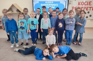 Grupa dzieci ubranych na niebiesko stoi obok siebie. Przed nimi dwoje dzieci leży, a jedna dziewczynka kuca. Za dziećmi jest tablica z napisem autyzm.