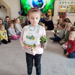 Dziewczynka stoi na szarym dywanie. W ręce trzyma doniczkę z roślinką, w drugiej trzyma dyplom za udział w konkursie ekologicznym. Na szyi wisi medal w kształcie roweru. Za dzieckiem jest ekran multimedialny z ilustracją ziemi trzymanej w rękach.