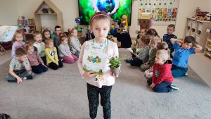 Dziewczynka stoi na szarym dywanie. W ręce trzyma doniczkę z roślinką, w drugiej trzyma dyplom za udział w konkursie ekologicznym. Na szyi wisi medal w kształcie roweru. Za dzieckiem jest ekran multimedialny z ilustracją ziemi trzymanej w rękach.