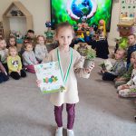 Dziewczynka stoi na szarym dywanie. W ręce trzyma doniczkę z roślinką, w drugiej trzyma dyplom za udział w konkursie ekologicznym. Na szyi wisi medal w kształcie roweru. Za dzieckiem jest ekran multimedialny z ilustracją ziemi trzymanej w rękach.