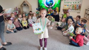 Dziewczynka stoi na szarym dywanie. W ręce trzyma doniczkę z roślinką, w drugiej trzyma dyplom za udział w konkursie ekologicznym. Na szyi wisi medal w kształcie roweru. Za dzieckiem jest ekran multimedialny z ilustracją ziemi trzymanej w rękach.