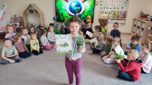 Dziewczynka stoi na szarym dywanie. W ręce trzyma doniczkę z roślinką, w drugiej trzyma dyplom za udział w konkursie ekologicznym. Na szyi wisi medal w kształcie roweru. Za dzieckiem jest ekran multimedialny z ilustracją ziemi trzymanej w rękach.