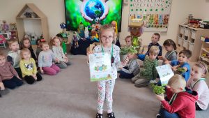 Dziewczynka stoi na szarym dywanie. W ręce trzyma doniczkę z roślinką, w drugiej trzyma dyplom za udział w konkursie ekologicznym. Na szyi wisi medal w kształcie roweru. Za dzieckiem jest ekran multimedialny z ilustracją ziemi trzymanej w rękach.