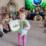 Dziewczynka stoi na szarym dywanie. W ręce trzyma doniczkę z roślinką, w drugiej trzyma dyplom za udział w konkursie ekologicznym. Na szyi wisi medal w kształcie roweru. Za dzieckiem jest ekran multimedialny z ilustracją ziemi trzymanej w rękach.