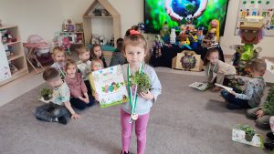 Dziewczynka stoi na szarym dywanie. W ręce trzyma doniczkę z roślinką, w drugiej trzyma dyplom za udział w konkursie ekologicznym. Na szyi wisi medal w kształcie roweru. Za dzieckiem jest ekran multimedialny z ilustracją ziemi trzymanej w rękach.