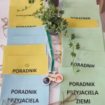 Na stole położone są obok siebie poradniki przyjaciela ziemi książeczka w kolorze niebieskim, żółtym, zielonym. Między nimi stoi roślinka w doniczce i medal w kształcie roweru.
